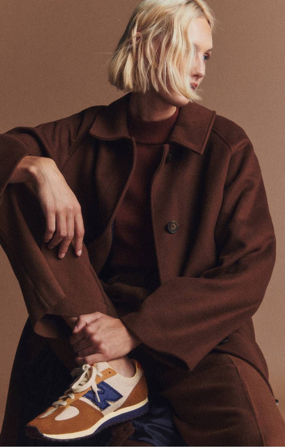 A woman sitting with a knee pulled up against a beige backdrop. 